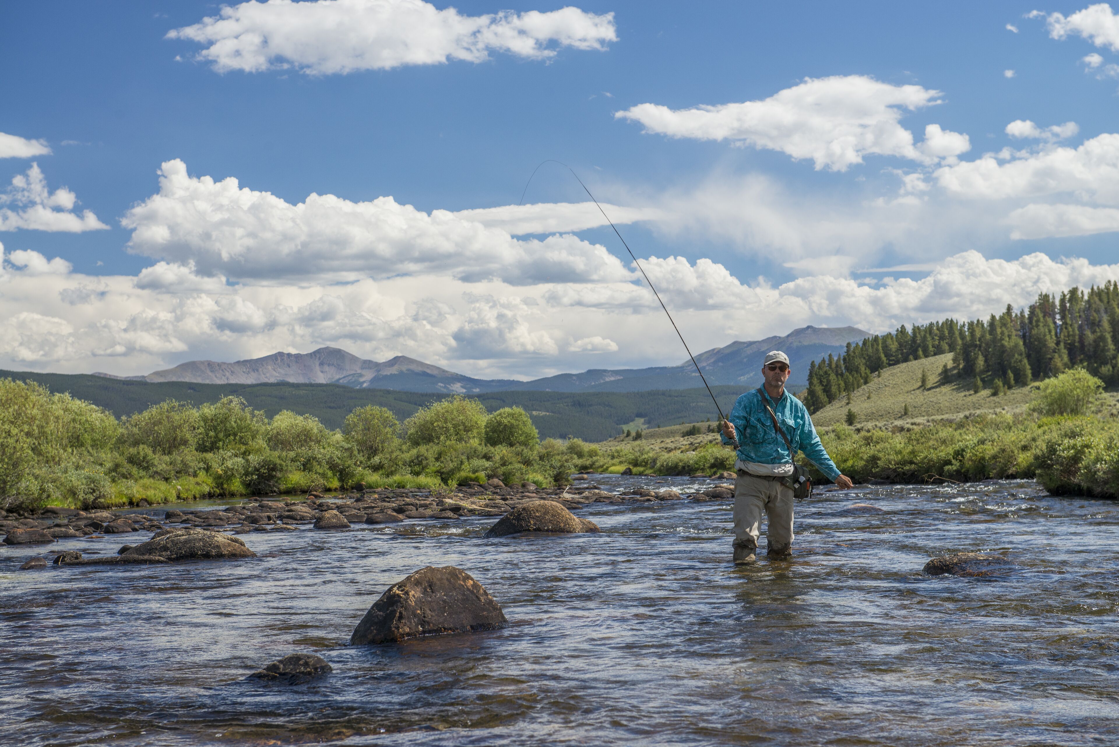 A person standing in a river with a fishing rod, surrounded by greenery and mountains in the background.