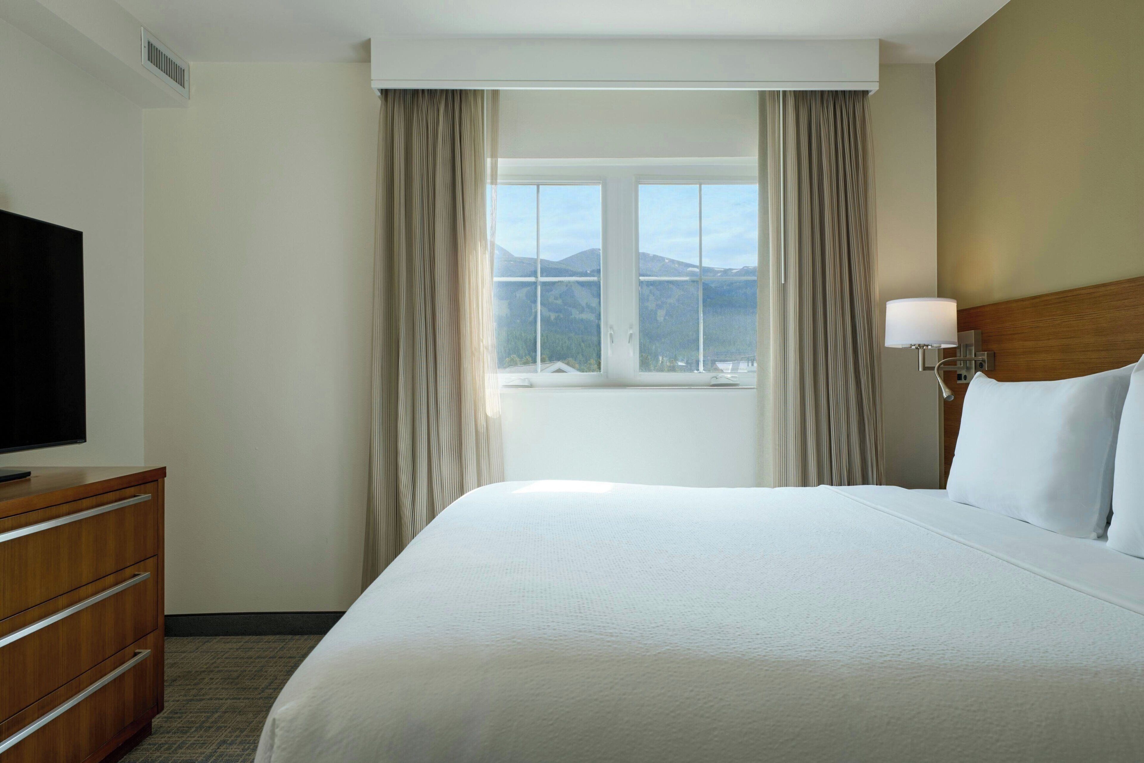 Une chambre d’hôtel avec un grand lit, un téléviseur posé sur une commode en bois, une fenêtre avec des rideaux et une lampe.