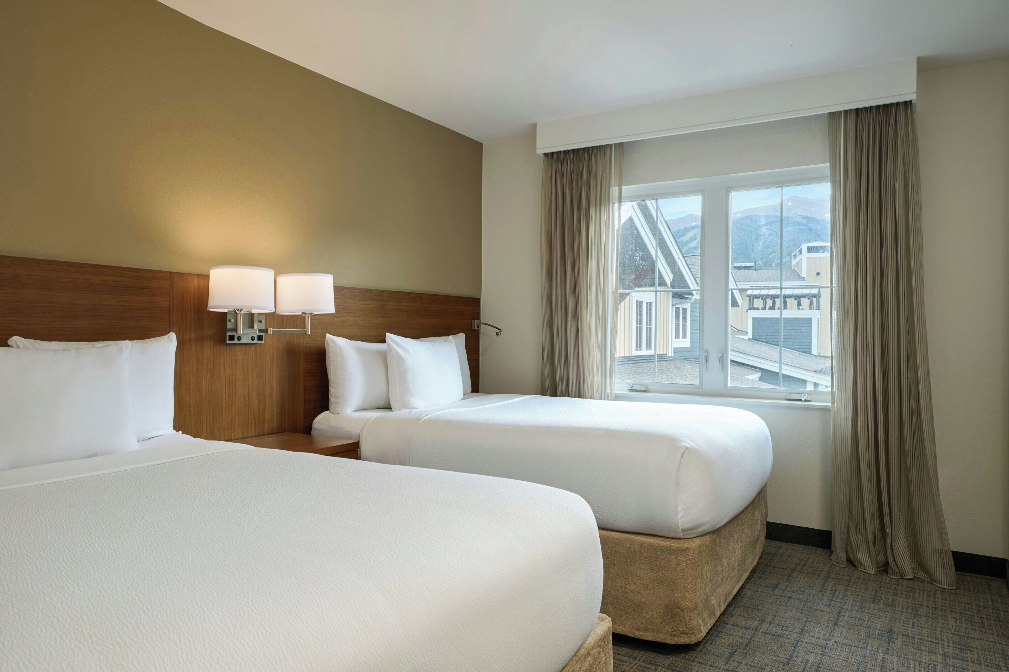 A hotel room with two beds, a wooden headboard, a nightstand with lamps, a window with curtains, and a view of buildings and mountains.