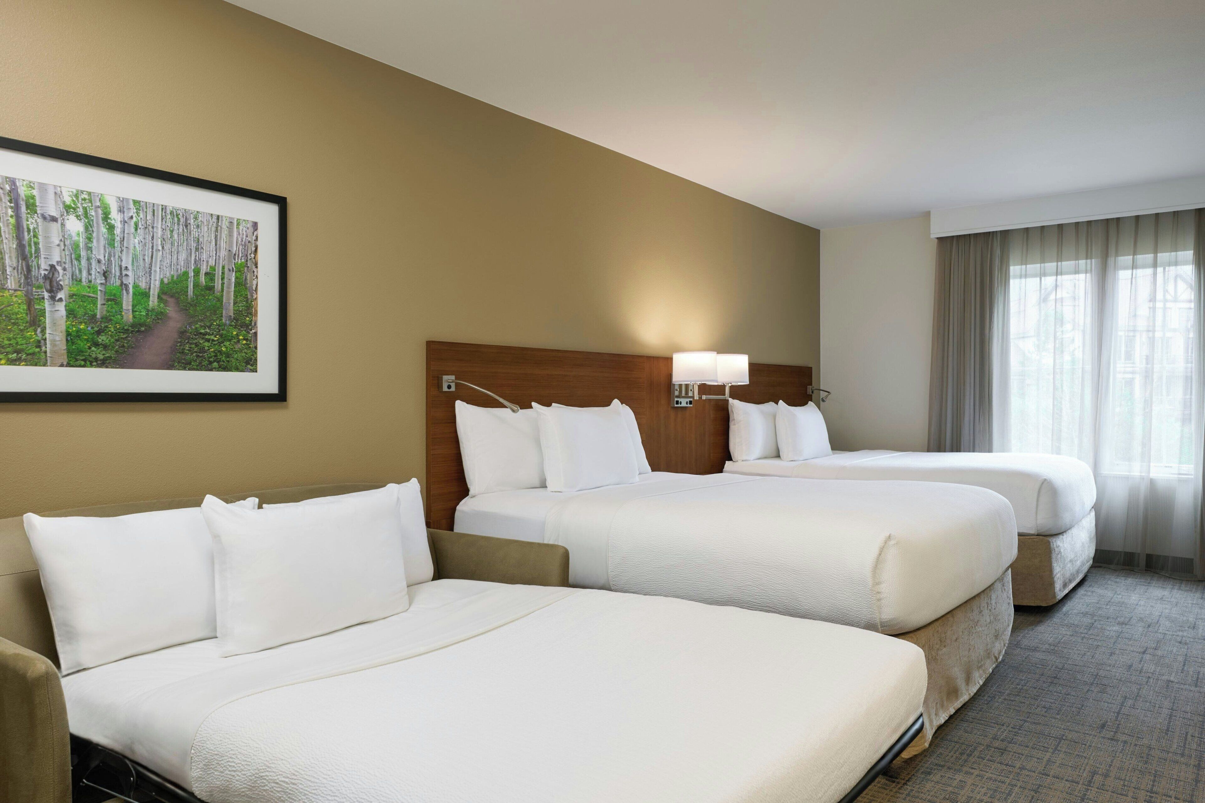 A hotel room with two beds, a headboard, a wall-mounted lamp, and a window with curtains.