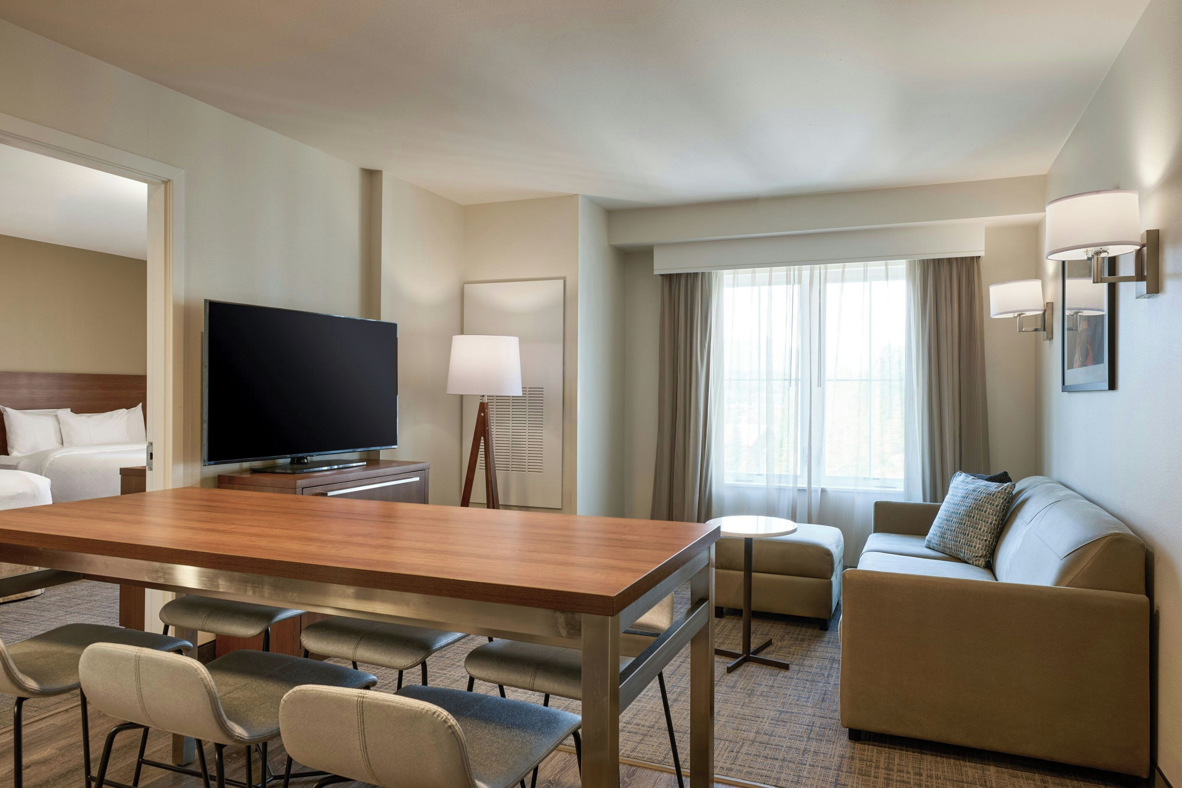 Une chambre d’hôtel équipée d’un téléviseur à écran plat, d’une table à manger en bois avec quatre chaises, d’un canapé, d’une petite table ronde et d’une grande fenêtre avec des rideaux.