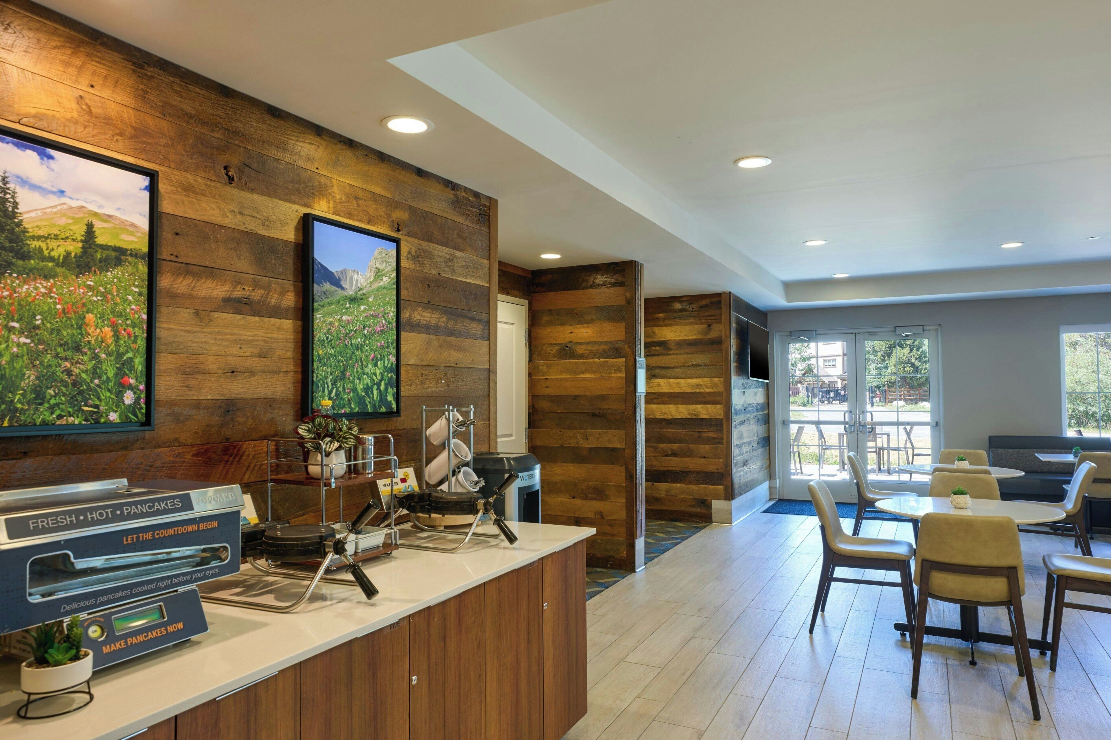 A modern hotel lobby with wooden paneling, a counter with a coffee machine, and a seating area with tables and chairs.