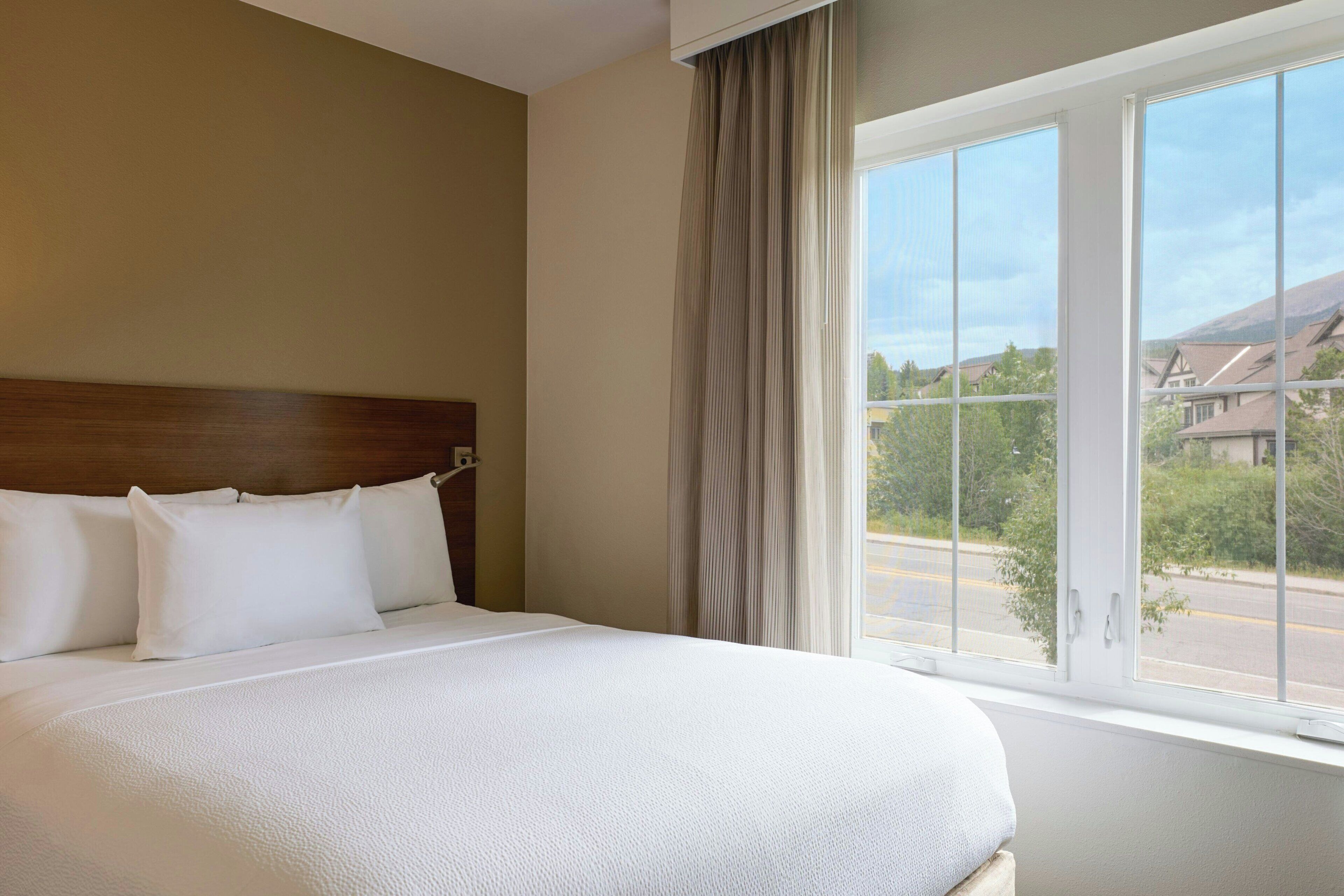 A hotel room with a large bed, a wooden headboard, and a window with a view of houses and greenery.