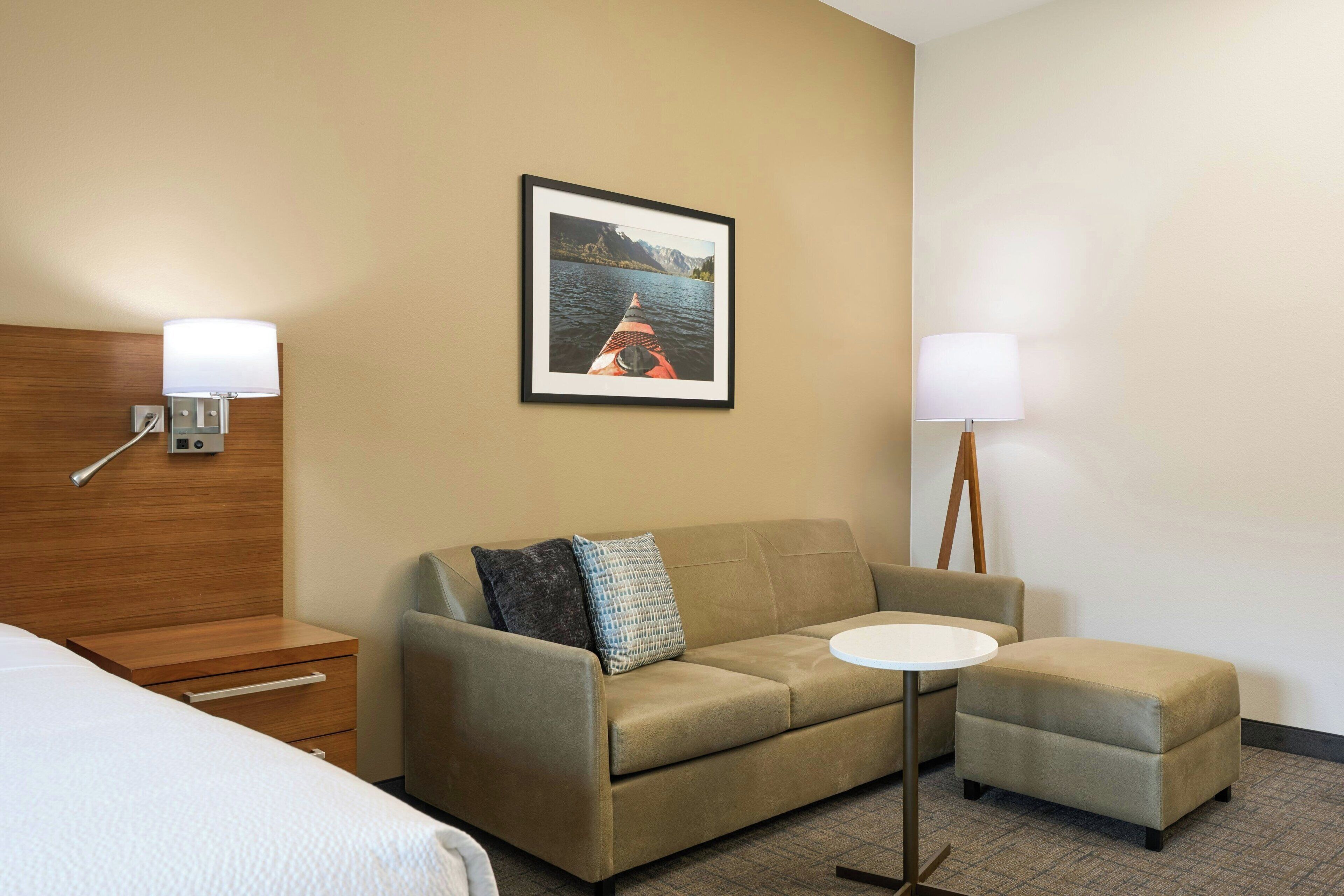 A hotel room with a sofa, an ottoman, a small table, a bed, and a framed picture on the wall.