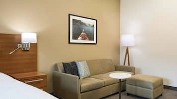 A hotel room with a sofa, an ottoman, a small table, a bed, and a framed picture on the wall.