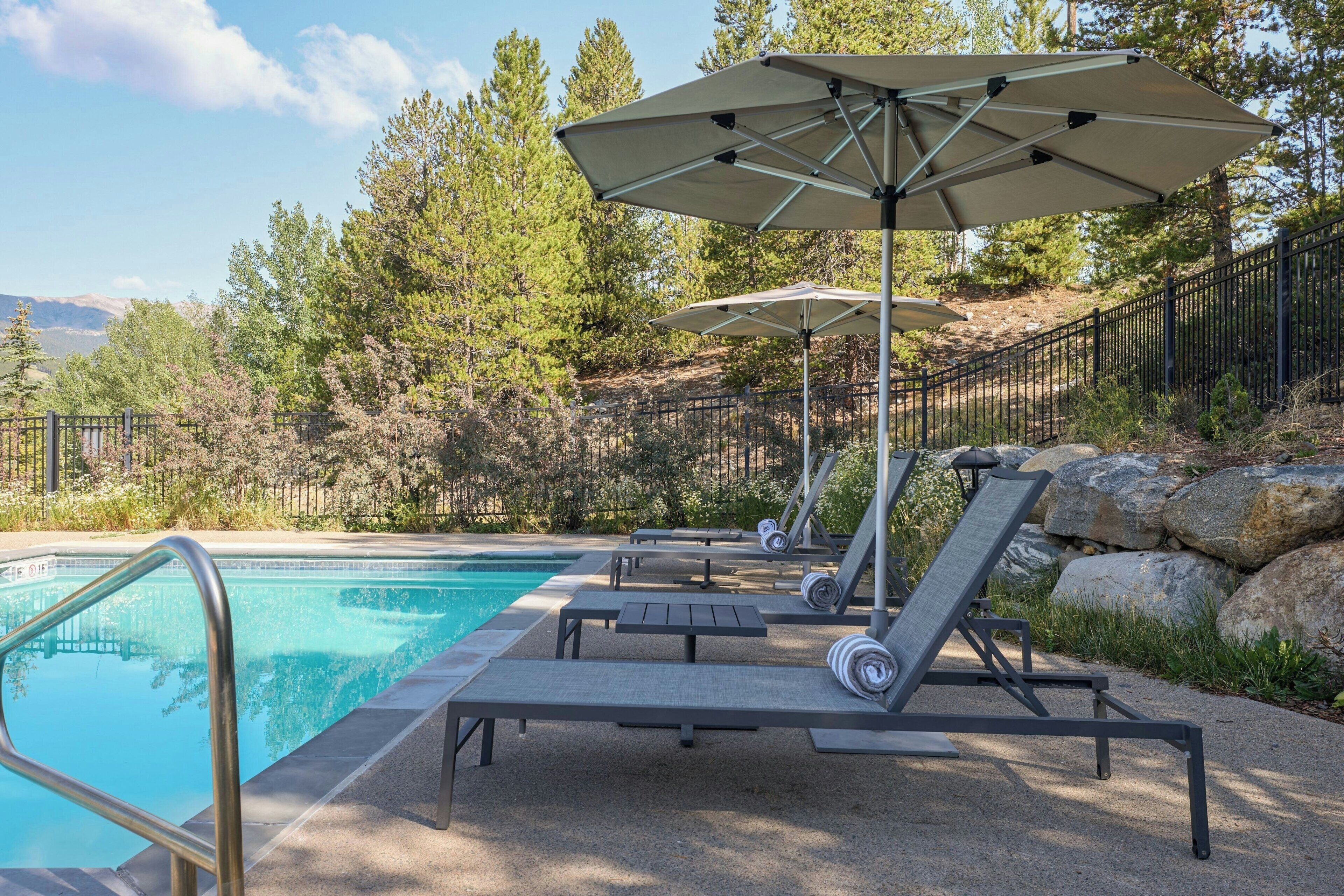 A poolside area with lounge chairs and umbrellas, surrounded by trees and a mountain view.