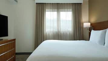 Una habitación de hotel con una cama grande, un televisor sobre una cómoda de madera, una ventana con cortinas y una mesita de noche con una lámpara.