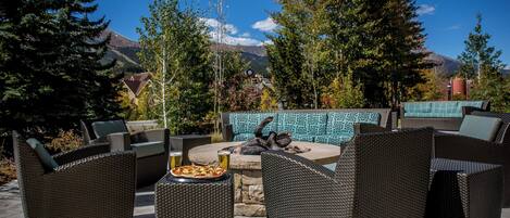 Un espace extérieur aménagé avec des meubles en osier, une table en pierre sur laquelle est posée une pizza, et une vue sur les montagnes.