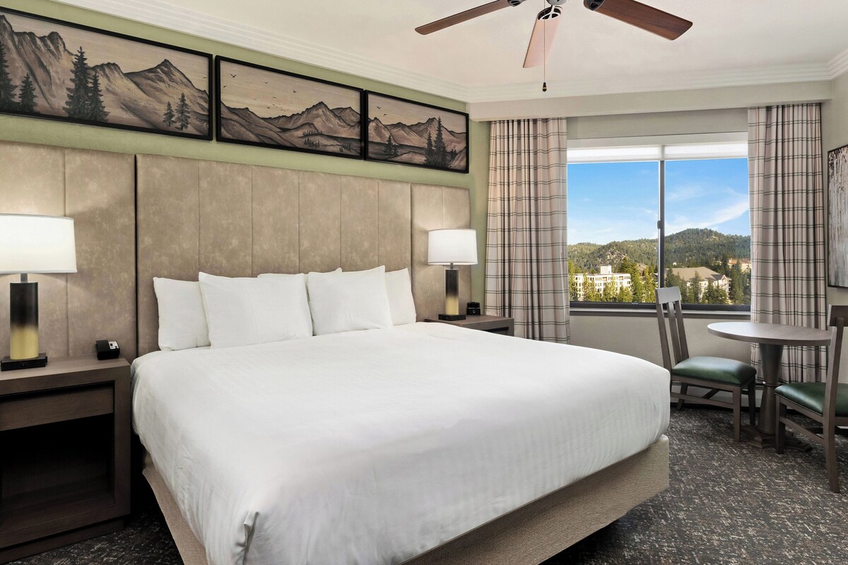 A hotel room with a large bed, two bedside lamps, a desk with a chair, a window with a view of mountains, and a ceiling fan.