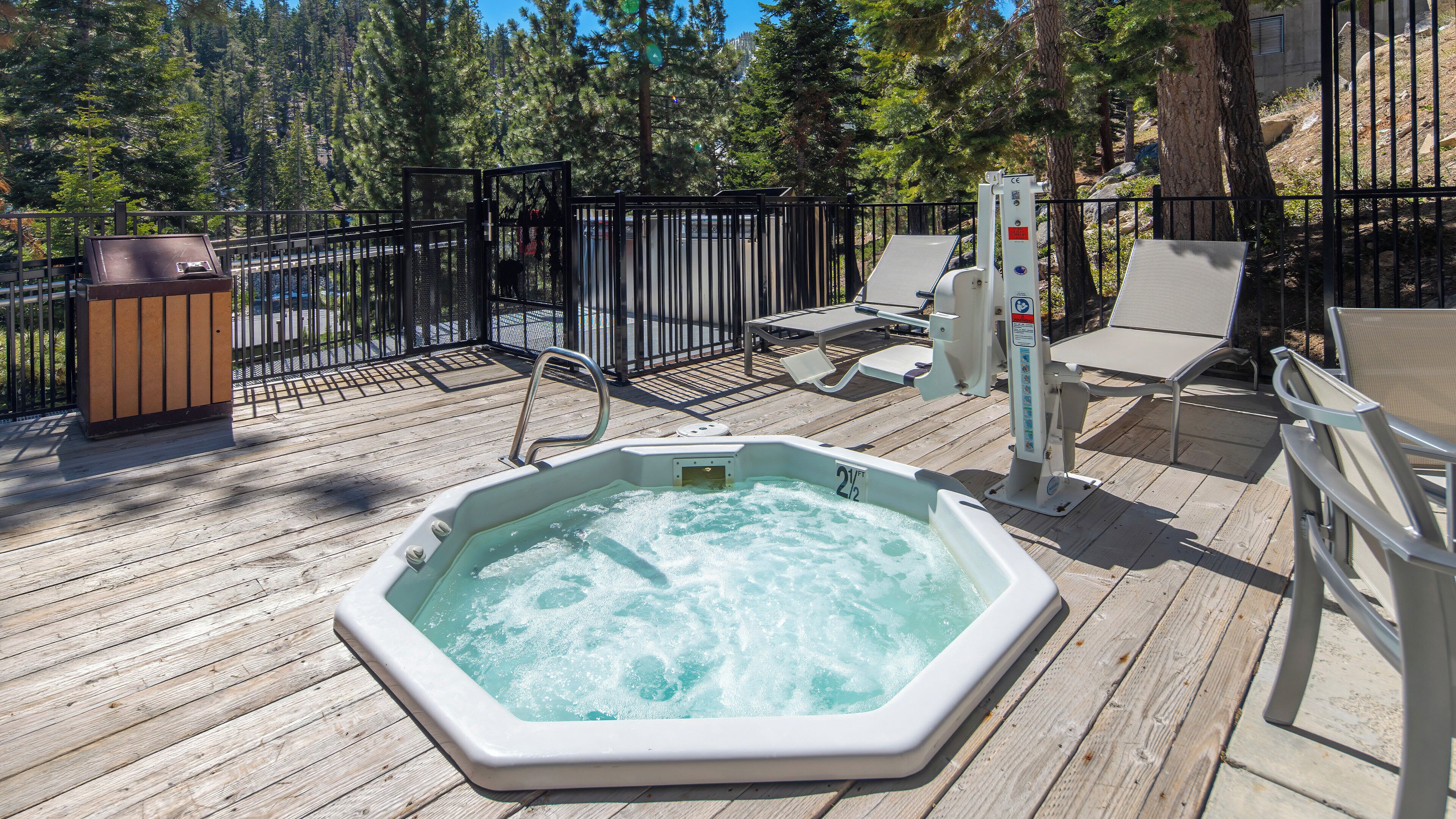 A hot tub on a wooden deck with lounge chairs and a fenced area.