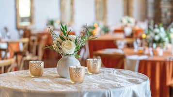 Una mesa redonda con mantel blanco, un florero con flores blancas y tres candelabros con textura.