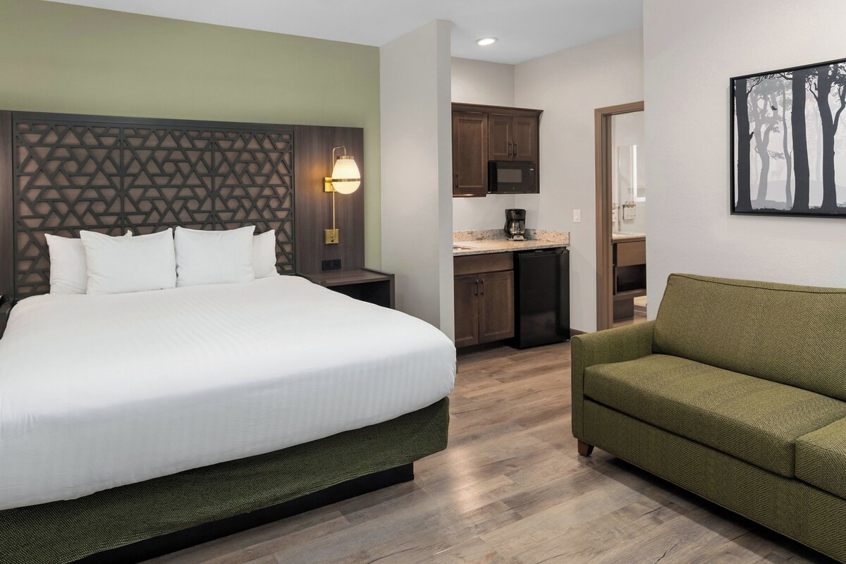 Quarto de hotel com cama, mesas de cabeceira, sofá verde, cozinha e quadro na parede.