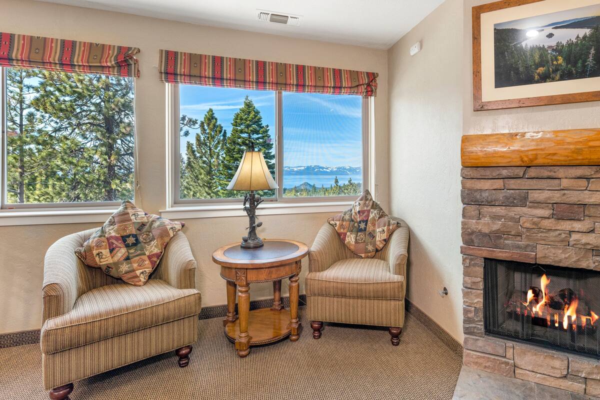 A cozy living room with two armchairs, a small round table with a lamp, a fireplace, and a large window with a view of trees and mountains.