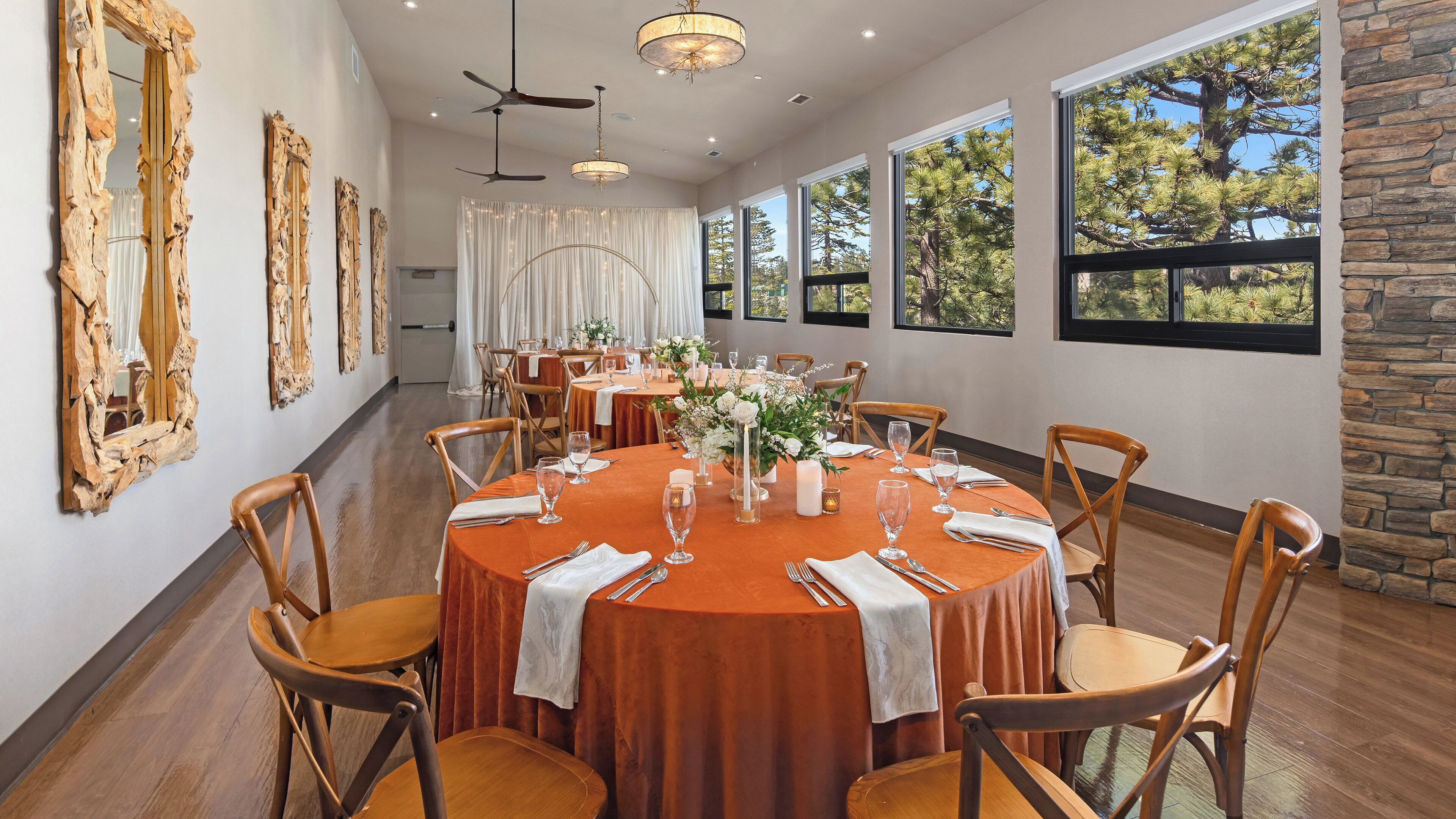 A dining room with round tables, wooden chairs, and large windows with a view of trees.