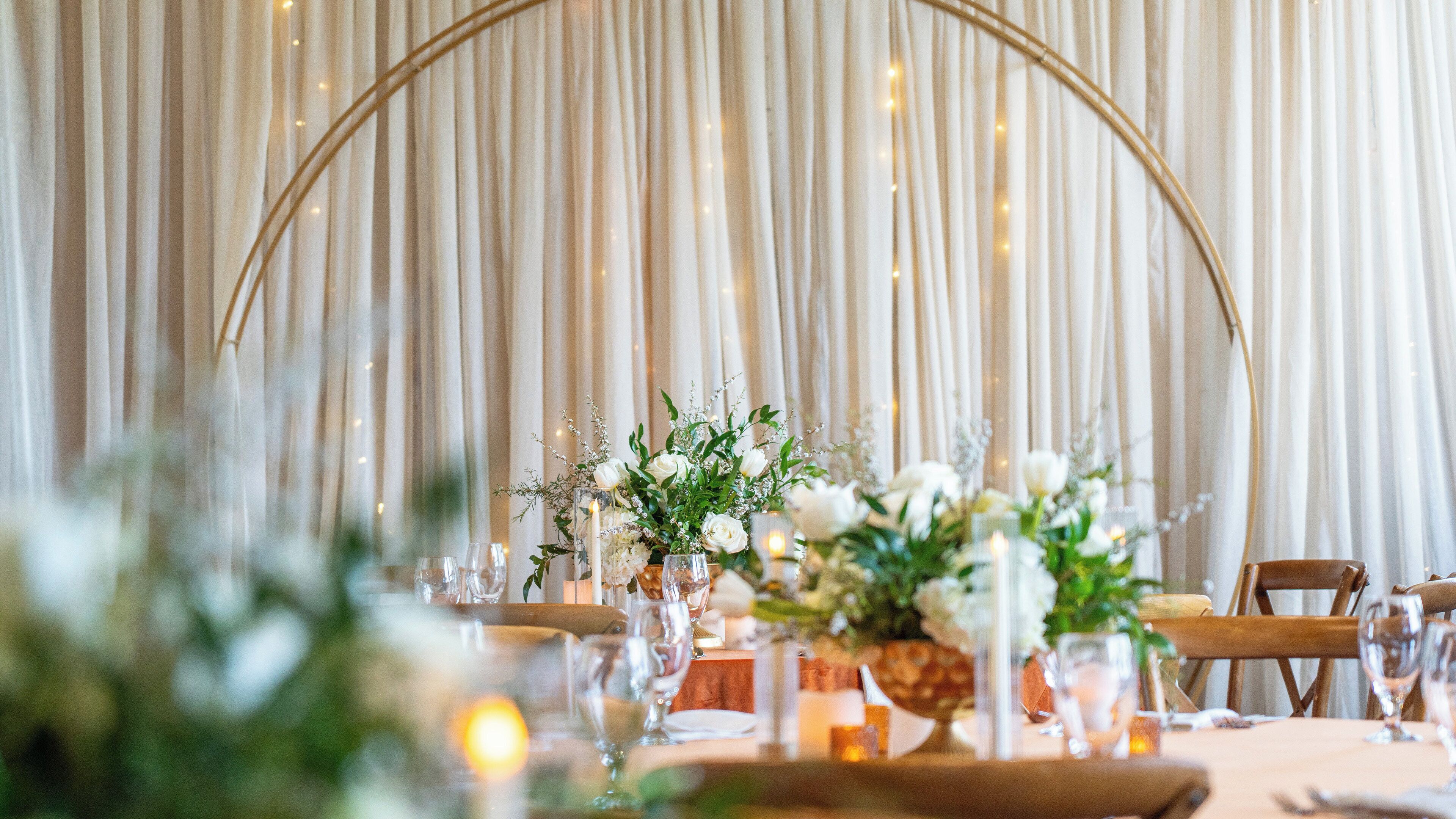 Una mesa dispuesta para un evento formal, con cortinas adornadas de fondo y centros de mesa florales.