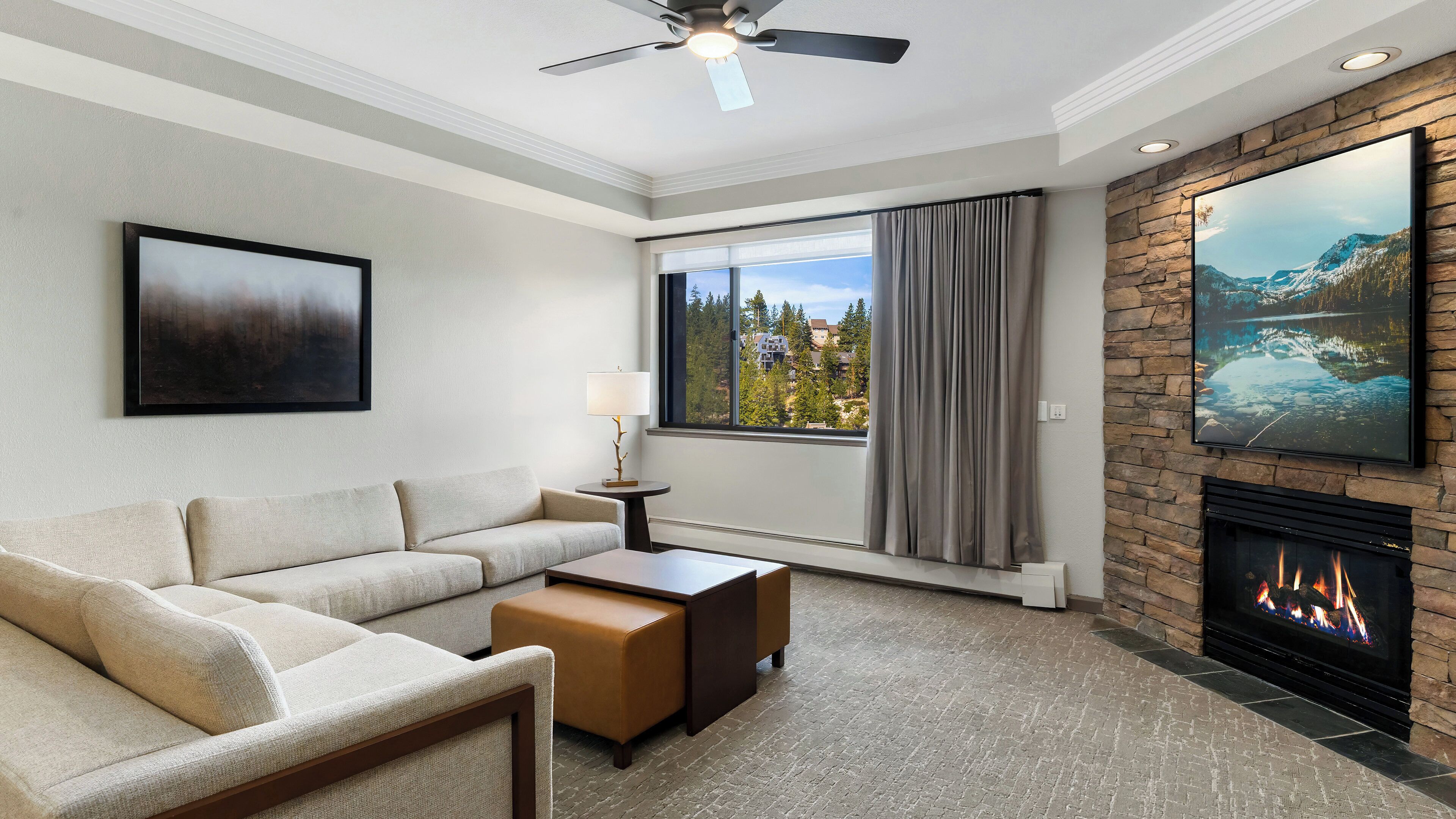 A modern living room with a sofa, a coffee table, a fireplace, a large window with a view, and a ceiling fan.
