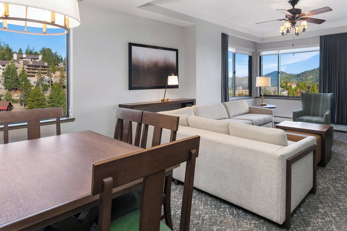 A modern hotel room with a dining area, a sofa set, a fireplace, and a large window with a mountain view.