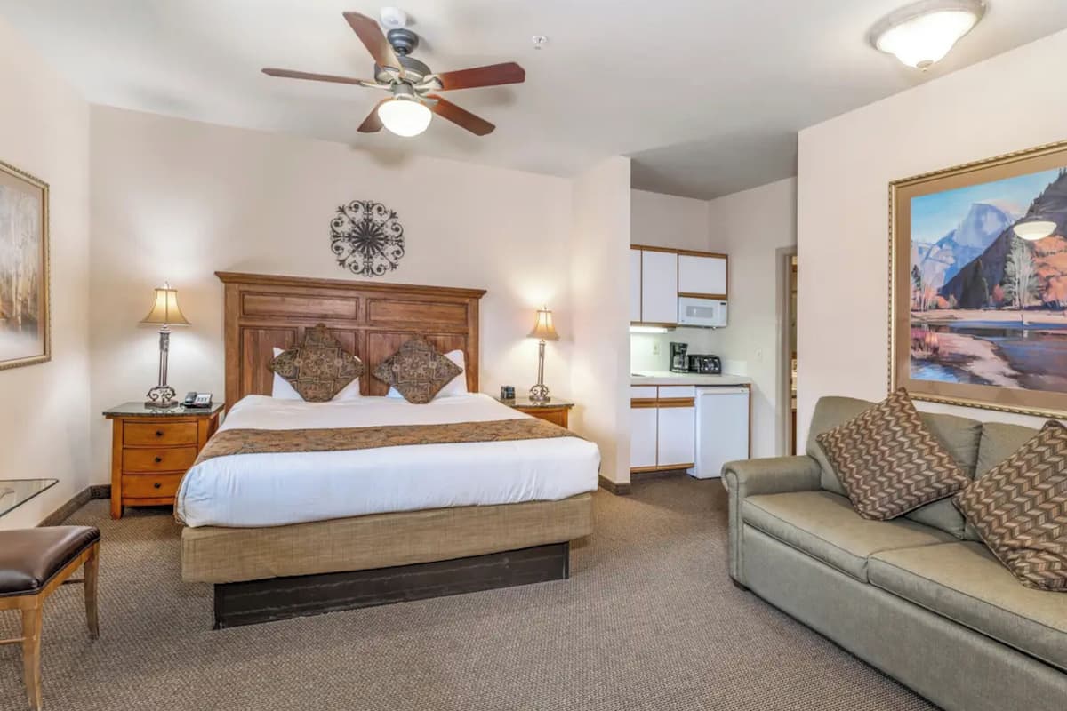 A hotel room with a bed, a desk, a chair, a sofa, a ceiling fan, a kitchen area, and a painting on the wall.