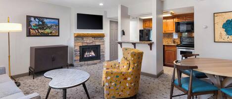 A living room with a fireplace, a television, a dining area with a table and chairs, and a kitchen area with cabinets and appliances.