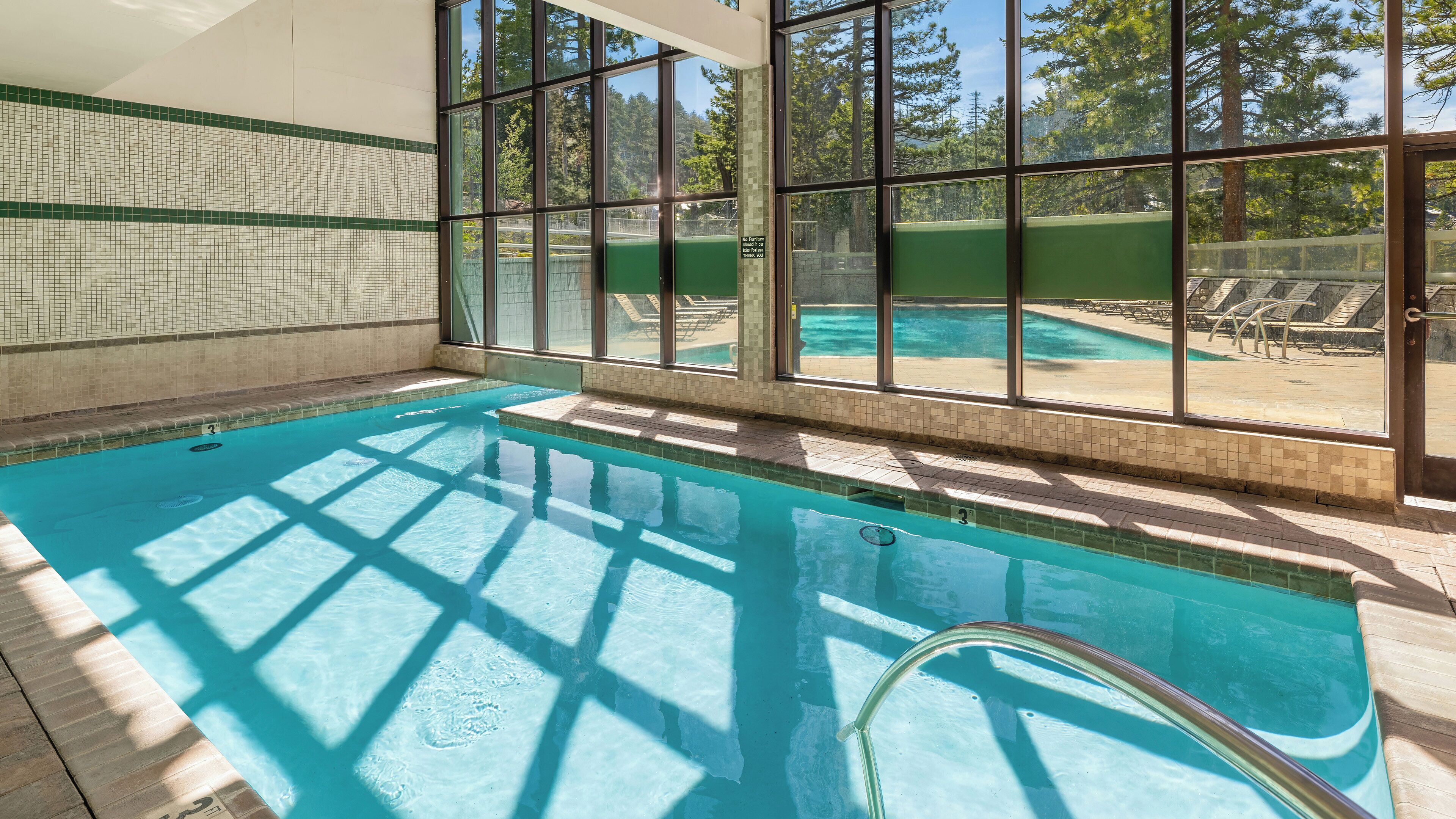 Una piscina interior con paredes de vidrio transparente, suelo de baldosas y vistas a una zona de piscina exterior.