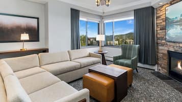 A hotel room with a sofa, armchair, coffee table, lamp, fireplace, and large windows with a view.