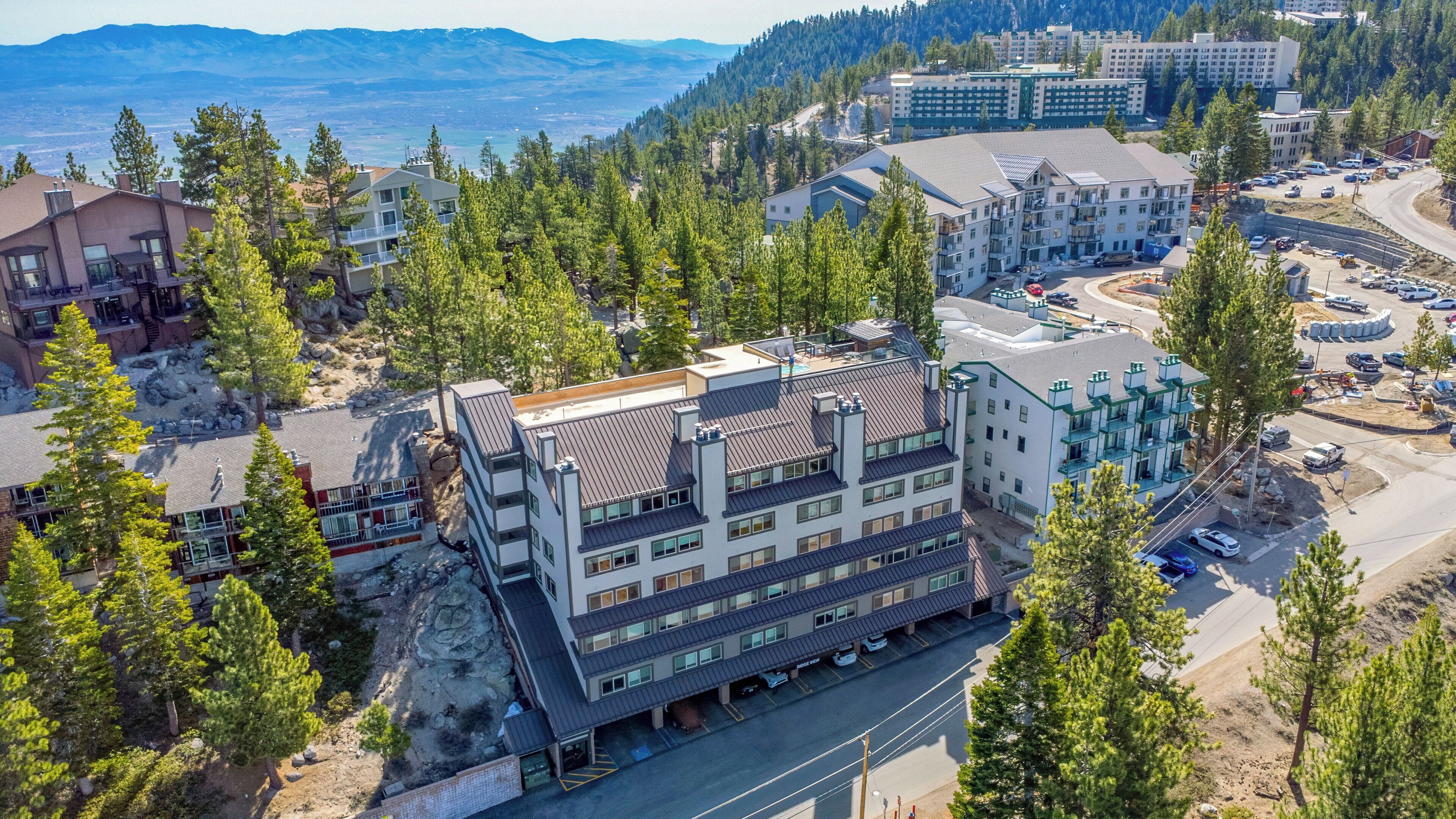 Un immeuble à plusieurs étages doté d’un parking, entouré d’arbres et avec une chaîne de montagnes en arrière-plan.