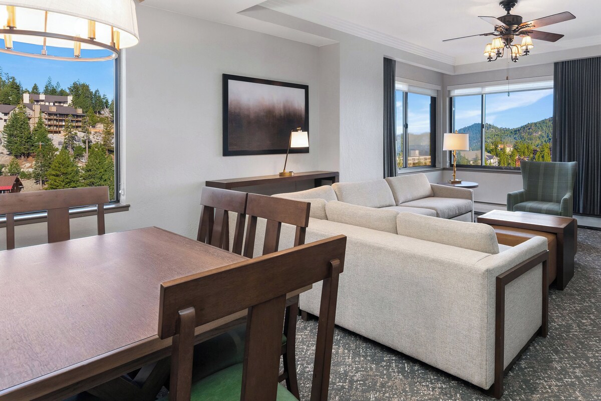 A modern hotel room with a dining area, a sofa set, a fireplace, and a large window with a mountain view.