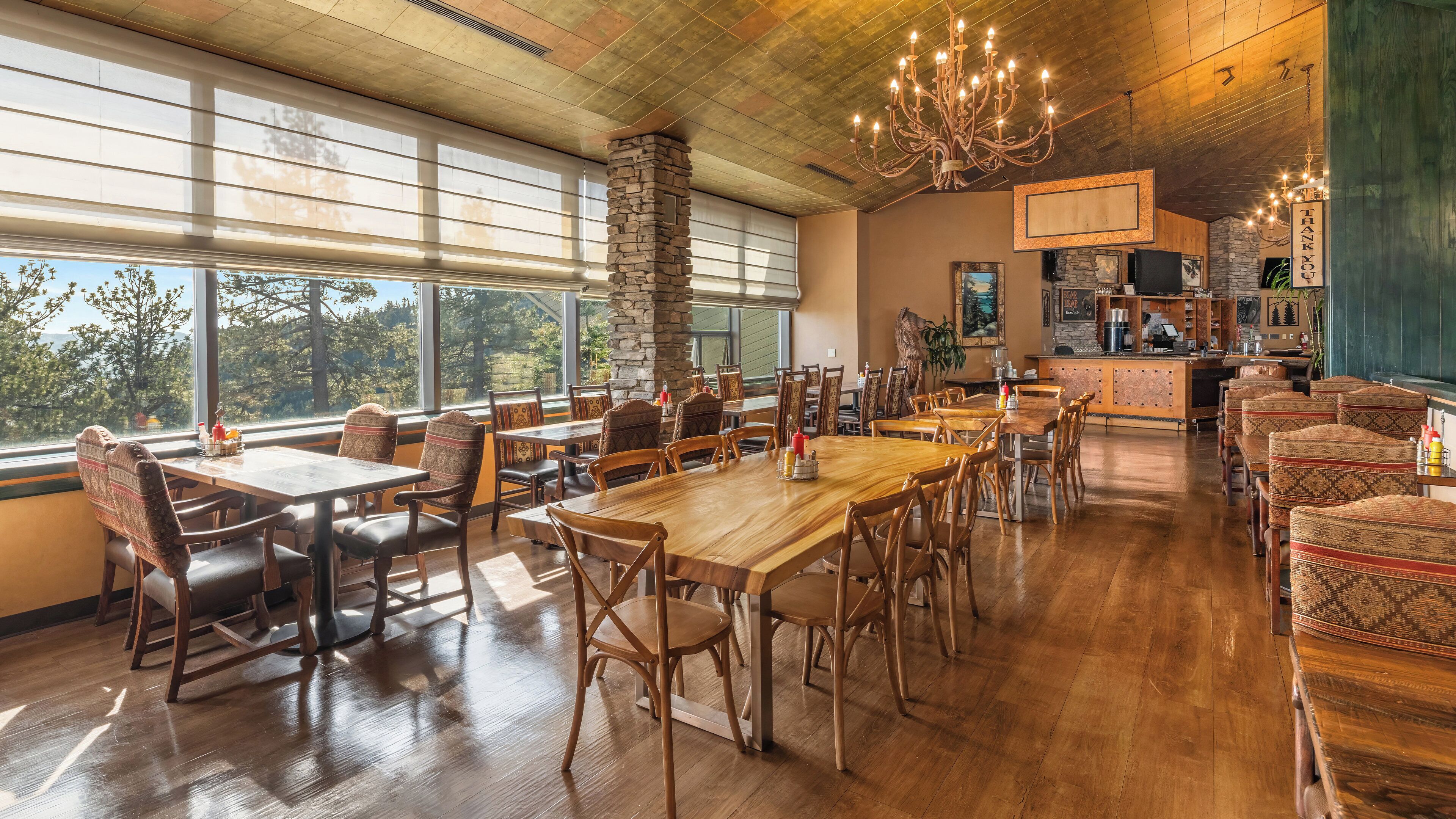 A spacious dining area with wooden tables and chairs, large windows with a view of trees, a stone pillar, and a chandelier.
