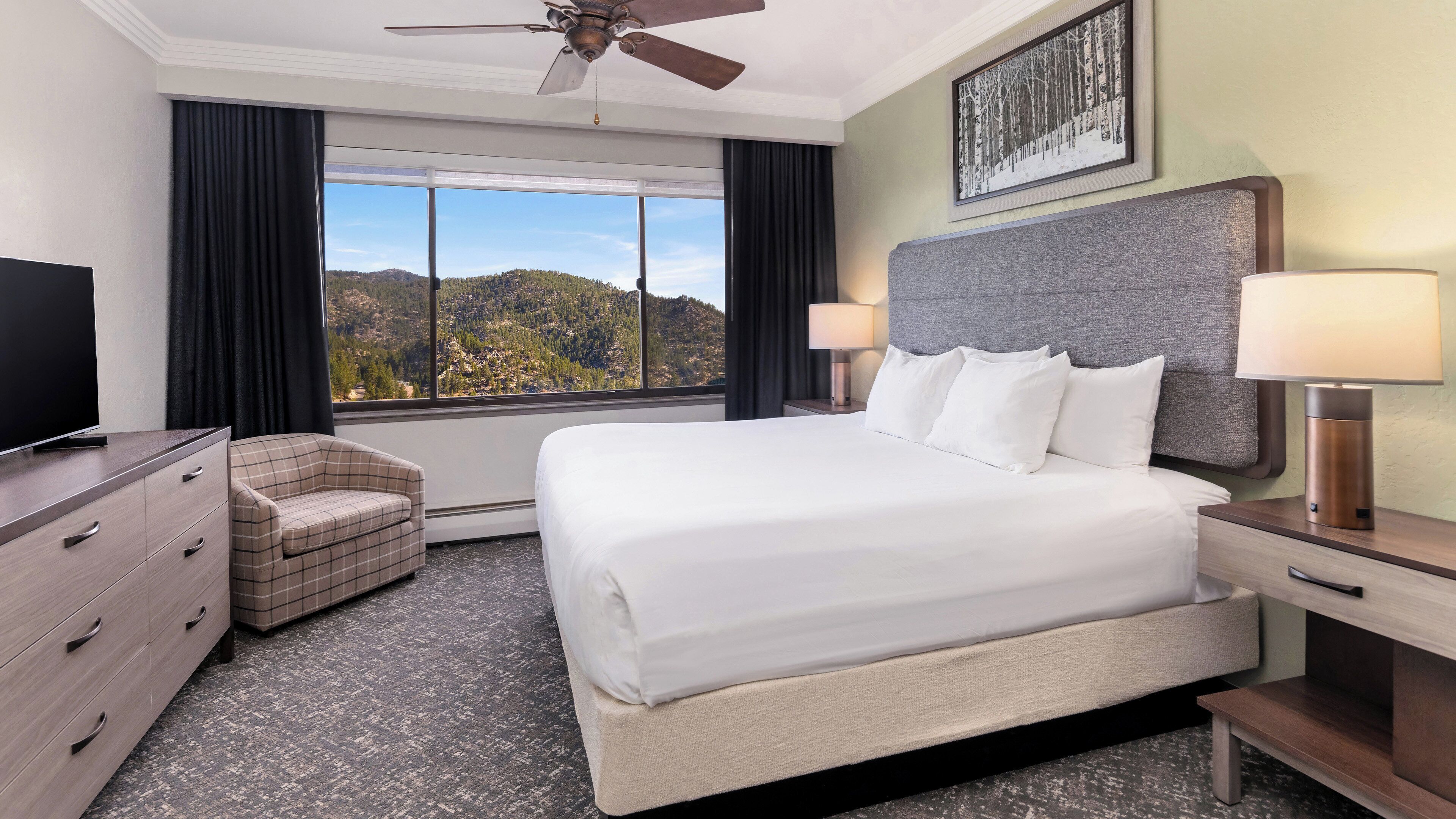 Ein Hotelzimmer mit Bett, Fernseher, Sessel, Schreibtisch mit Lampe und Blick auf eine gebirgige Landschaft.