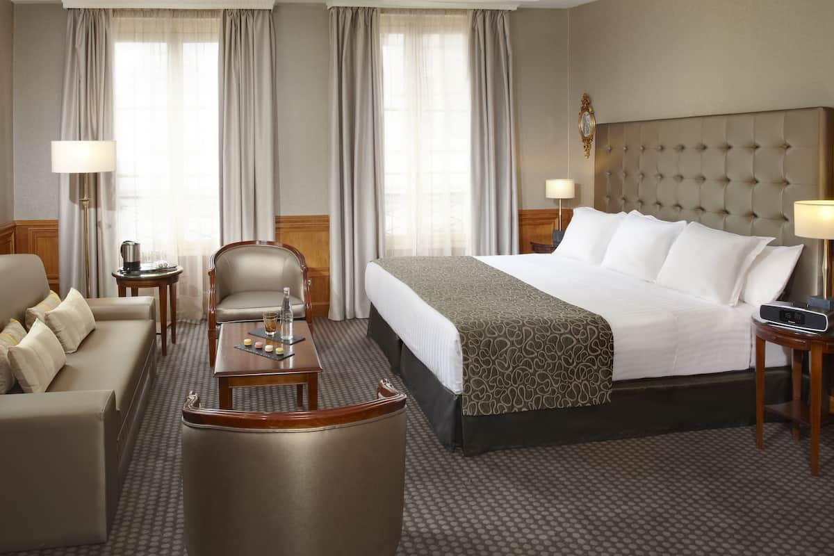 A hotel room with a bed, a sofa, a chair, a small table, and a bedside table with a lamp.