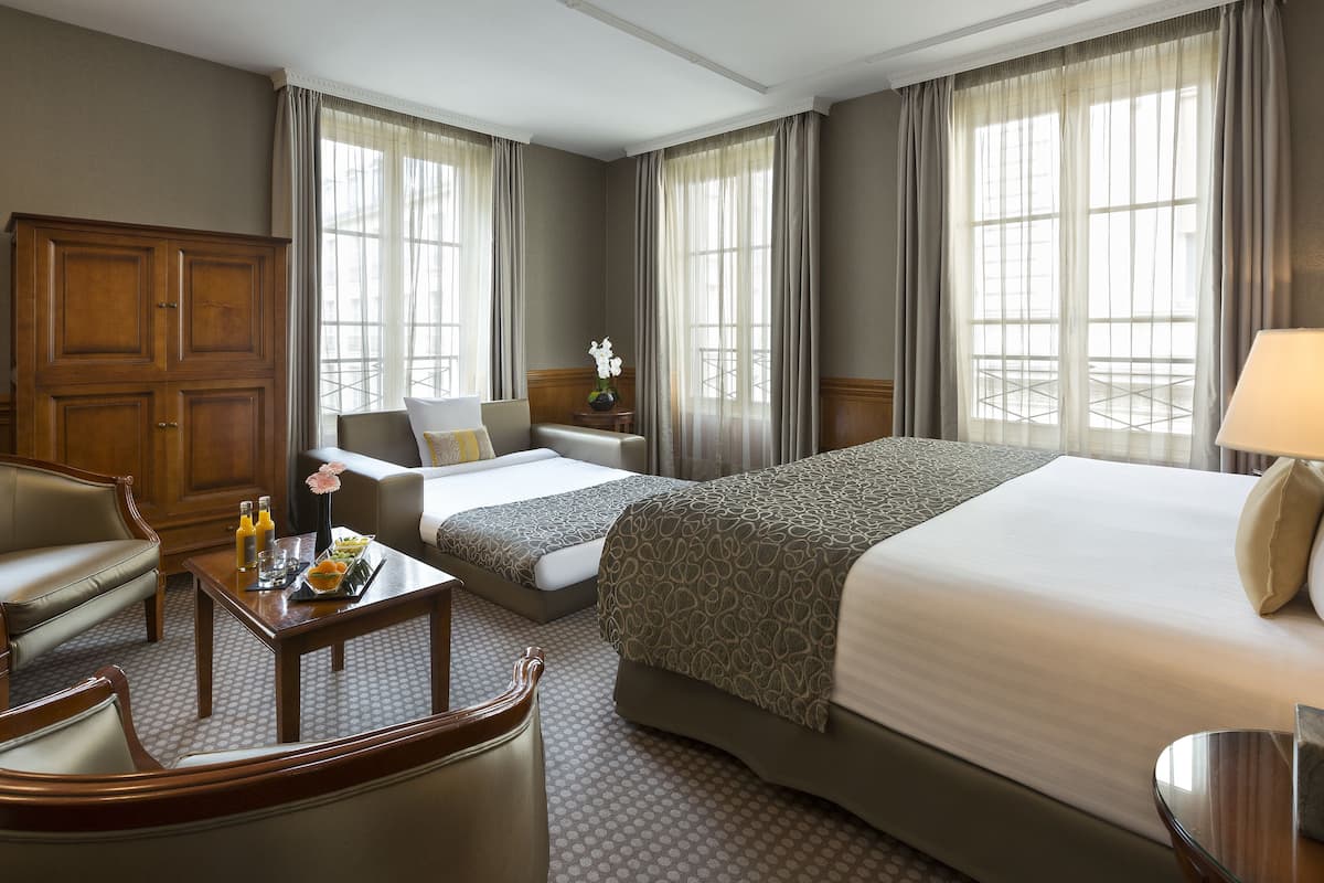 A hotel room with two beds, a sofa, a wooden cabinet, a small table with a tray of breakfast items, and large windows with curtains.