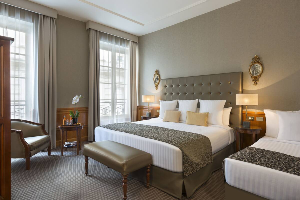A hotel room with two beds, a sitting area with a chair and bench, a small table with a vase of flowers, and a large window with curtains.