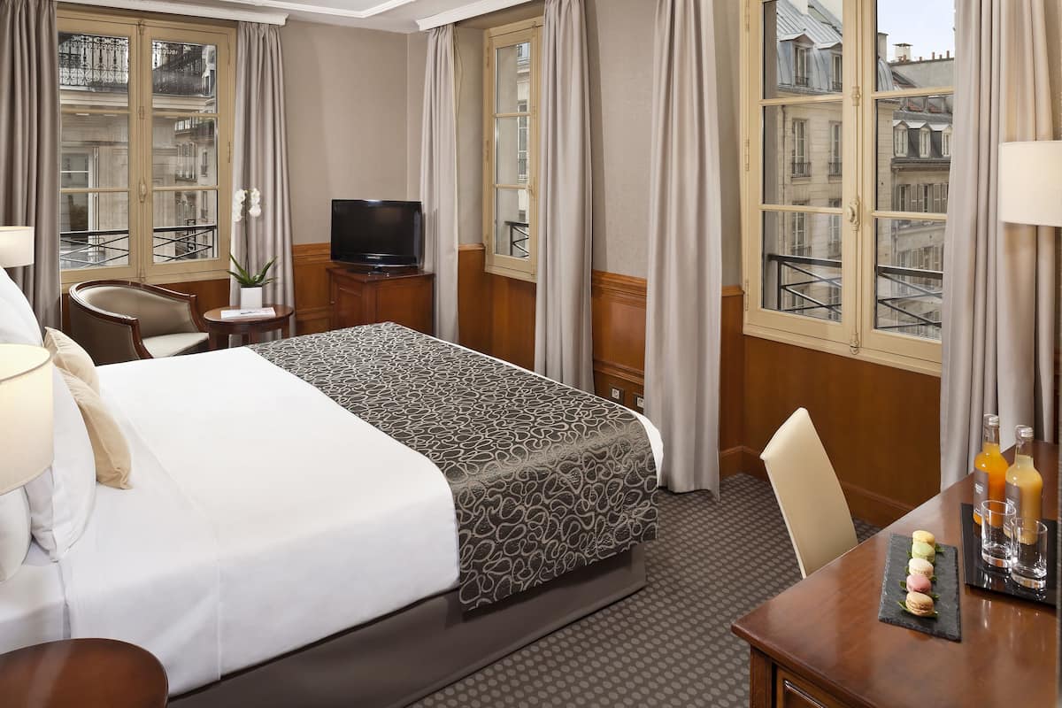 A hotel room with a large bed, a TV, a desk with a chair, and a view of the city through large windows.