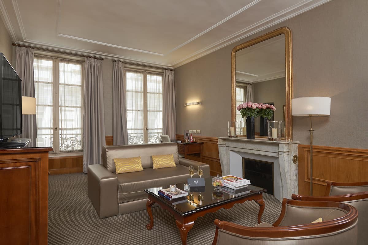 A well-appointed hotel room with a sofa, armchairs, a coffee table, a fireplace, and a large mirror.