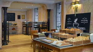 Un bufé de desayuno con diversos pasteles y pan, mesas y sillas de madera, y un menú en la pizarra en un ambiente de cafetería.