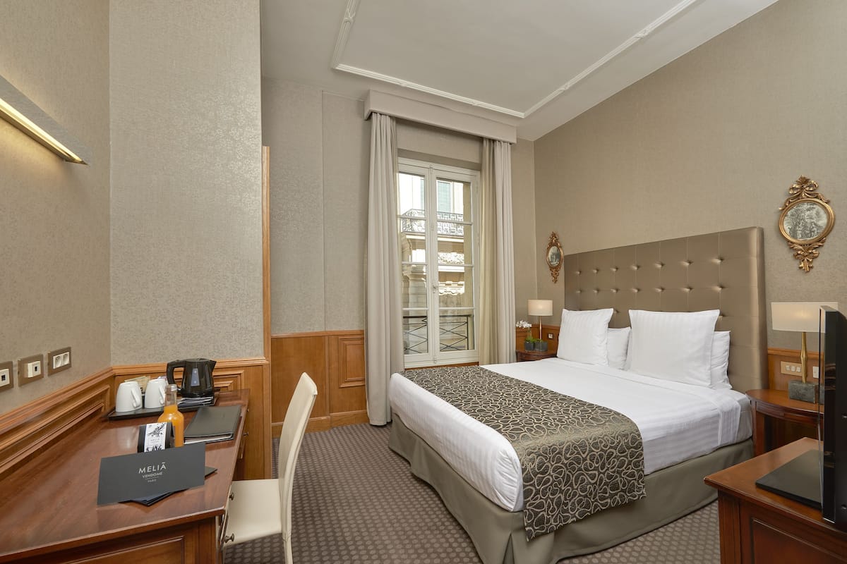 A hotel room with a large bed, a desk with a coffee maker, a chair, and a window with curtains.