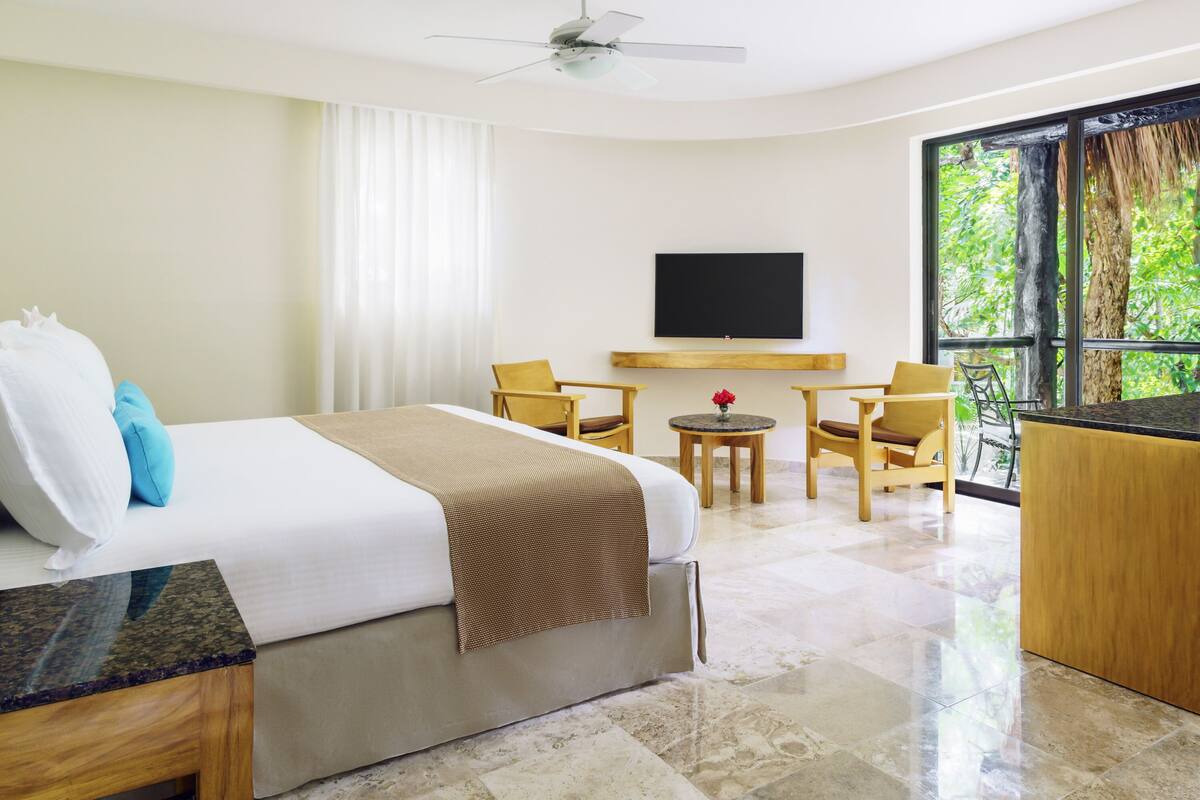 A hotel room with a bed, a TV, a seating area with chairs, a small table, and a large window with a view of trees.