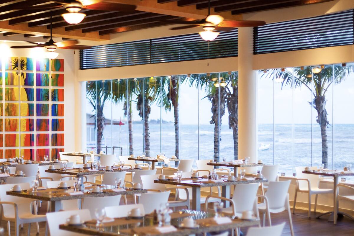 A restaurant with tables set for dining, large windows with a view of the ocean, and colorful abstract art on the walls.
