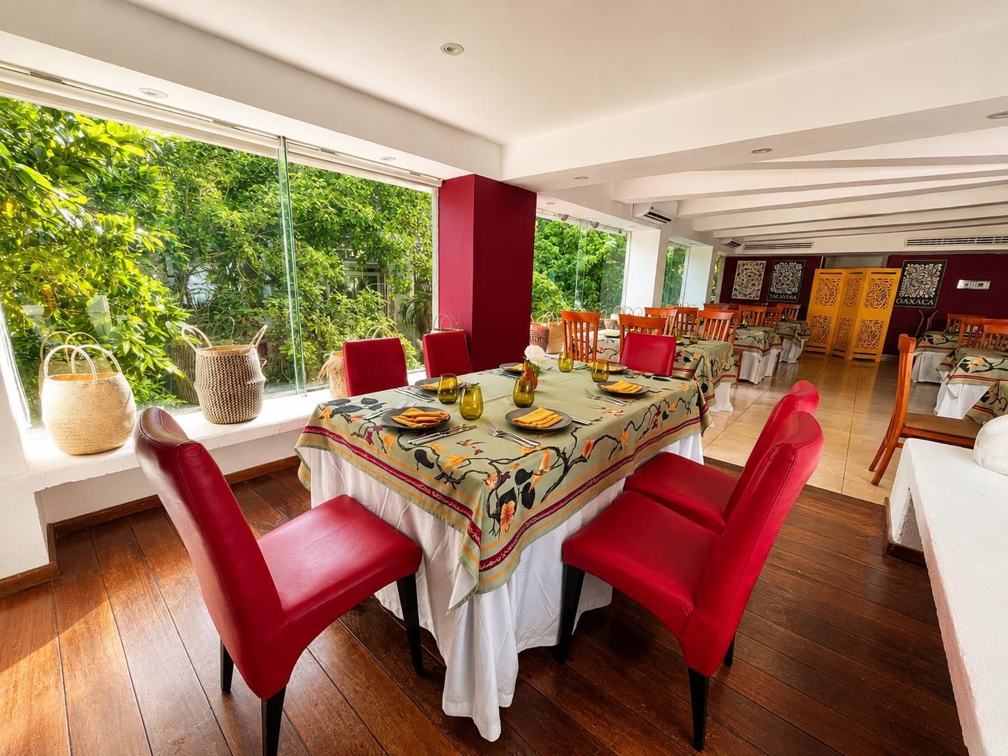 A dining area with a large table set for four, surrounded by red chairs, with a view of lush greenery outside.