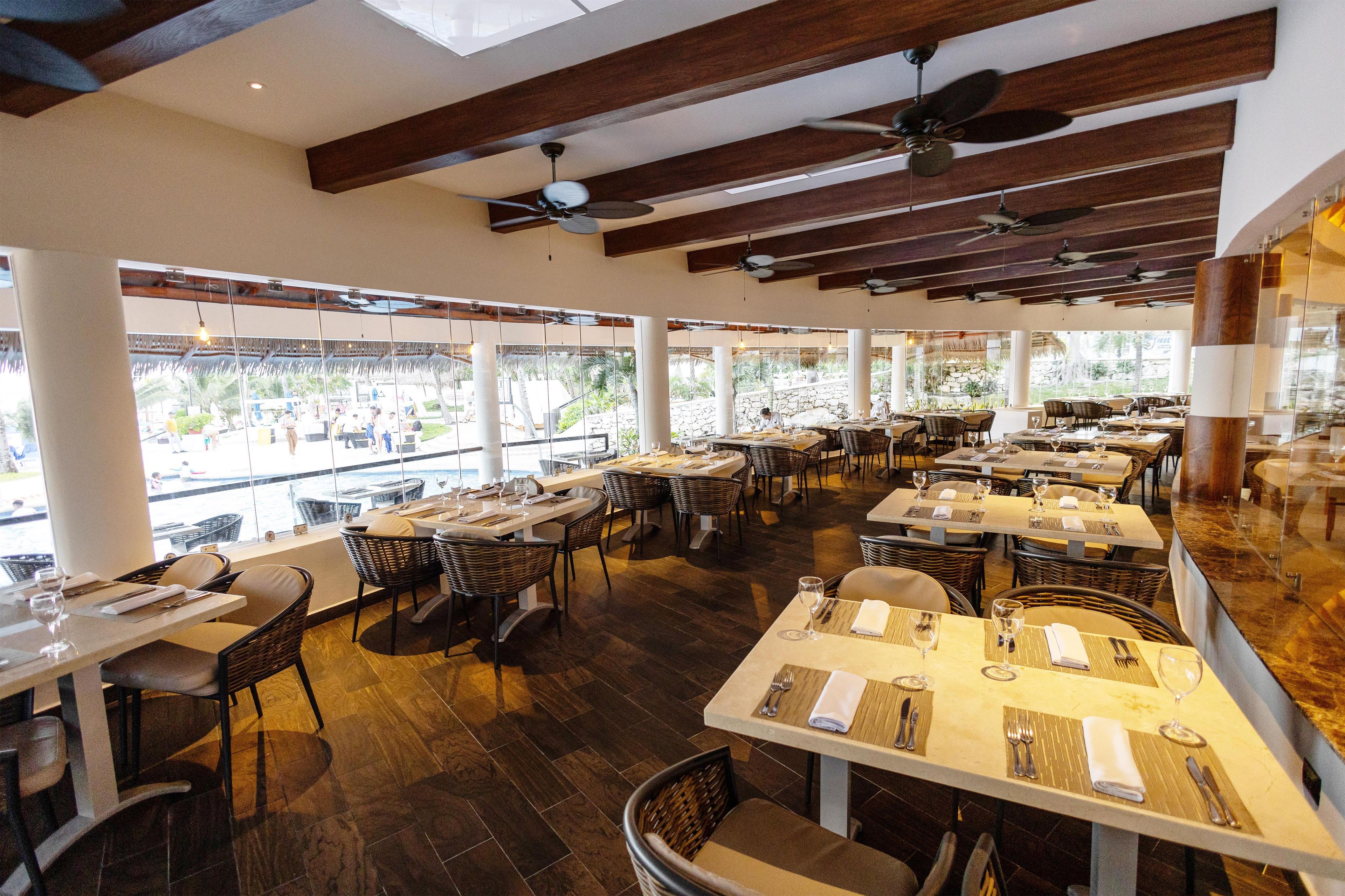 A restaurant with multiple dining tables and chairs, large windows offering a view of the outside, and ceiling fans.