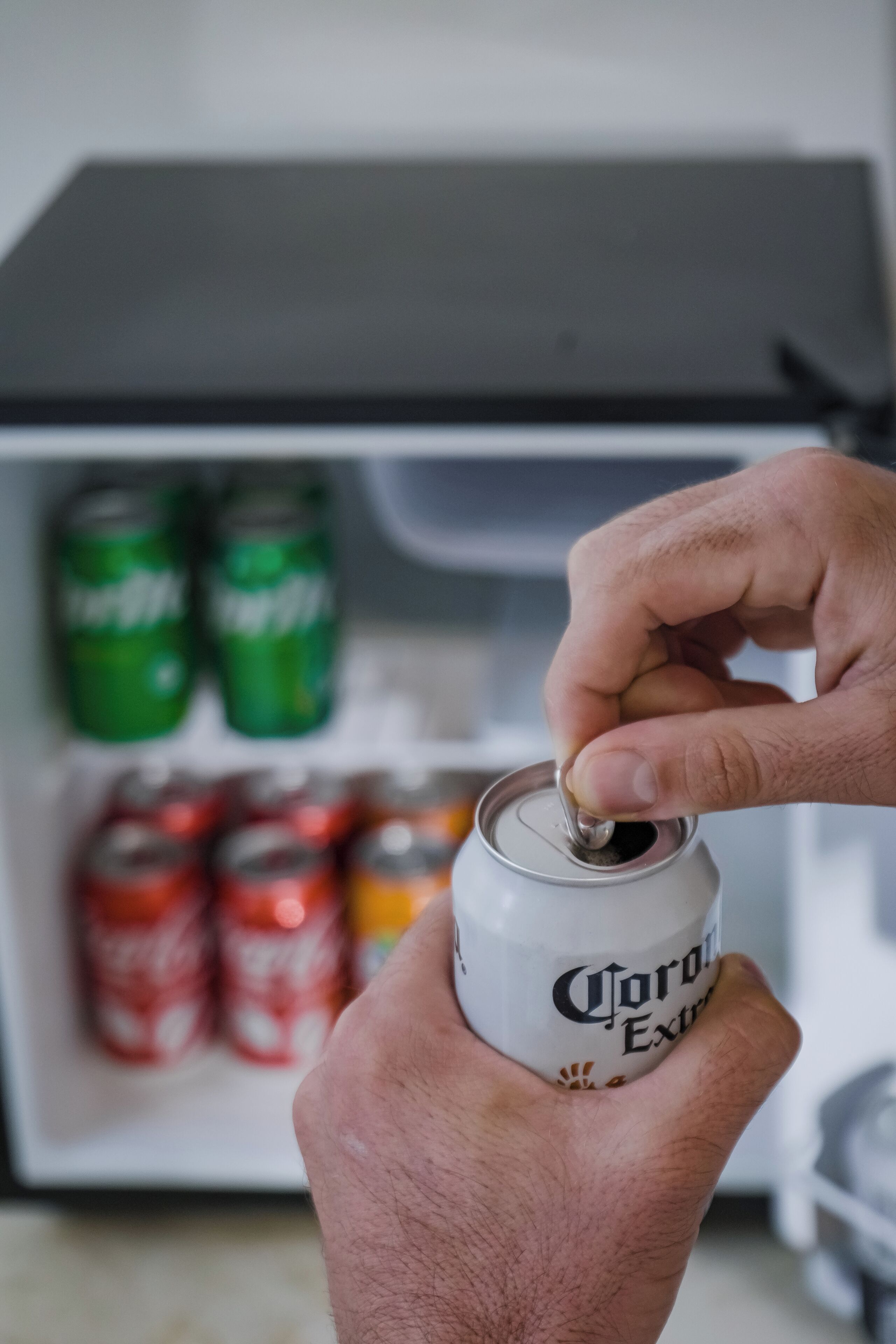 Una mano sostiene una lata de cerveza Corona Extra. Al fondo, se observan estantes con diversas latas de cerveza, incluyendo latas verdes y rojas.