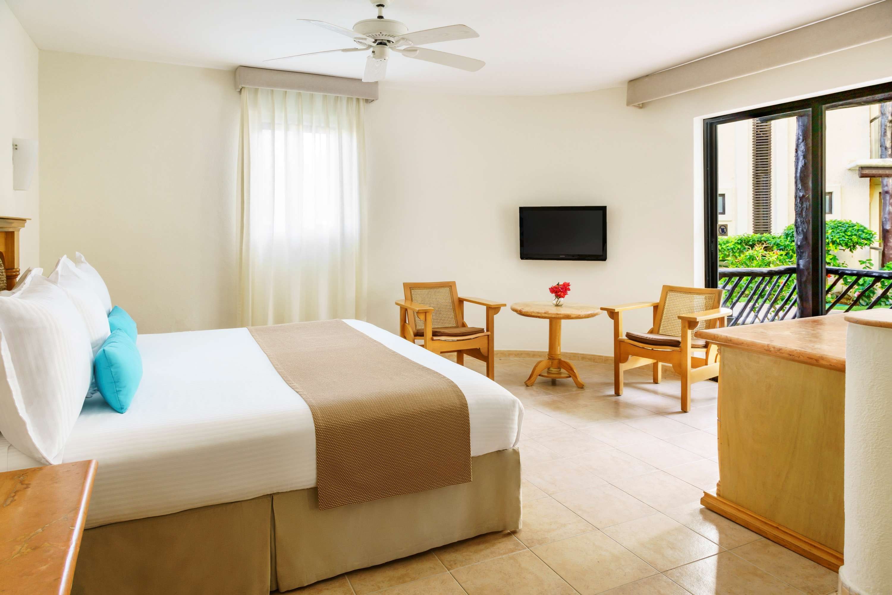A hotel room with a bed, a desk, a chair, a small table, a ceiling fan, and a TV on the wall.