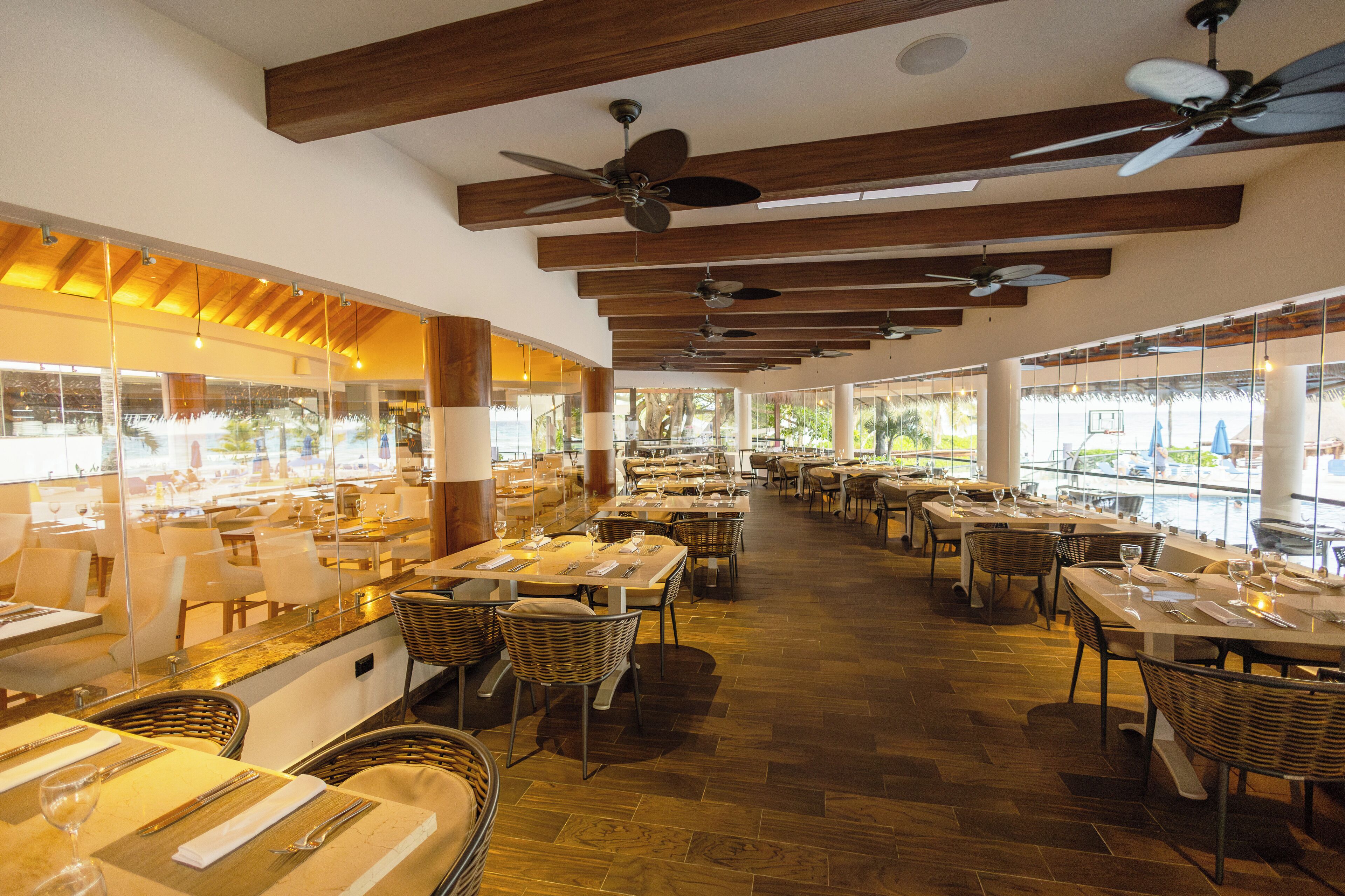 Un restaurant spacieux avec de hauts plafonds, des poutres en bois et de nombreux ventilateurs de plafond. Les tables sont recouvertes de nappes blanches et entourées de chaises en osier. De grandes fenêtres offrent une vue sur l’extérieur.