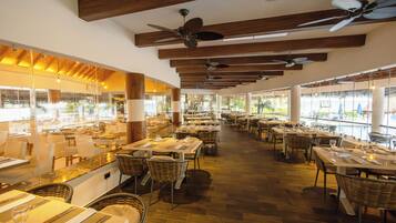 Un restaurante espacioso con techos altos, vigas de madera y varios ventiladores de techo. Las mesas están cubiertas con manteles blancos y sillas de mimbre. Amplias ventanas ofrecen vistas al exterior.