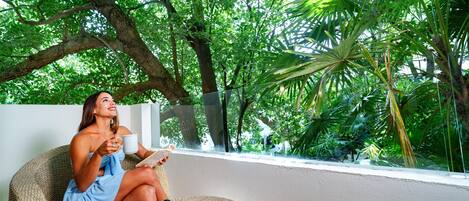 A person seated on a wicker chair with a book and a mug, surrounded by lush greenery and a white wall.