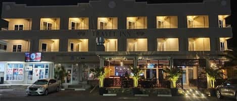 Un edificio de varios pisos con el letrero "TRAVEL INN", que indica que se trata de un hotel. Se observan varias ventanas en cada planta, y se aprecia la entrada con un área delimitada por una reja y plantas en macetas.