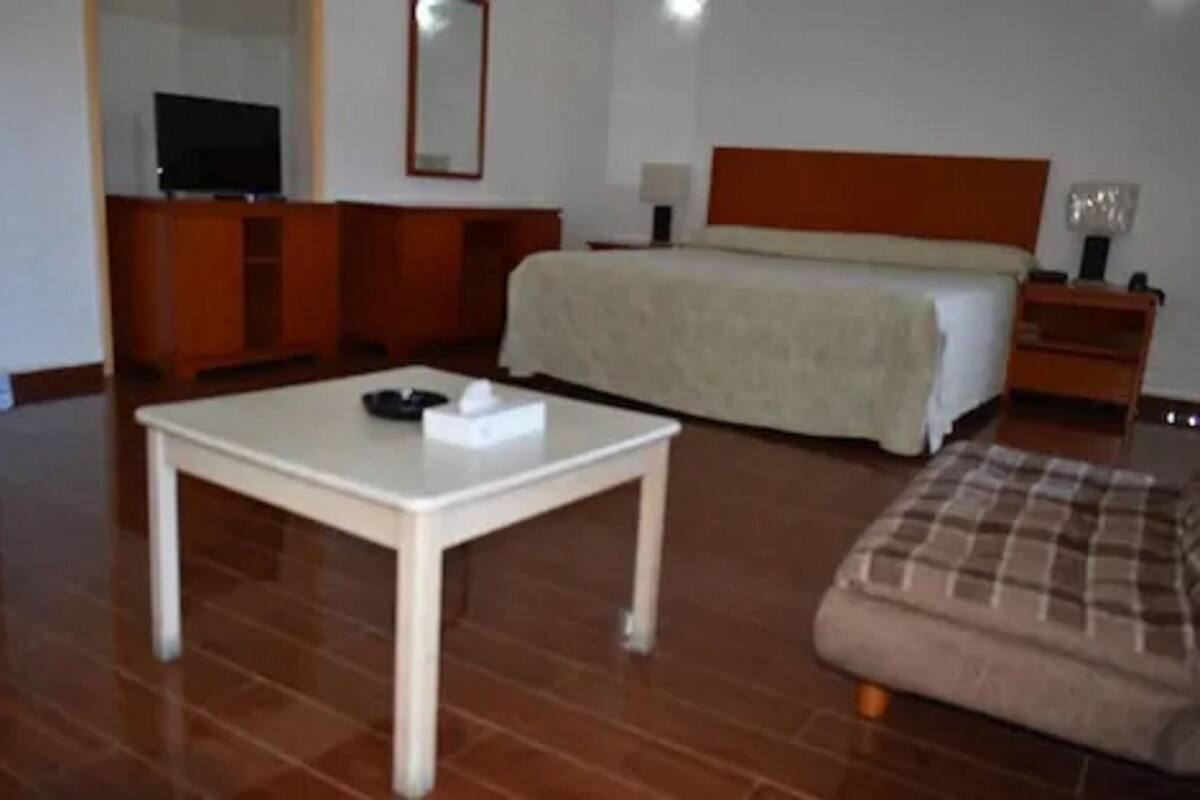 Quarto de hotel com uma cama, televisão em uma cômoda, mesa de cabeceira e uma pequena mesa com caixa de lenços.