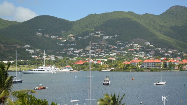A marina with various boats, including sailboats and a yacht, surrounded by lush green hills and a coastal town with red-roofed buildings. - Travel Inn Hotel (Simpson Bay)