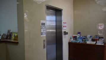 Un ascenseur dans le hall d’un hôtel, avec une affiche sur le mur et un comptoir en bois où sont disposées des brochures.