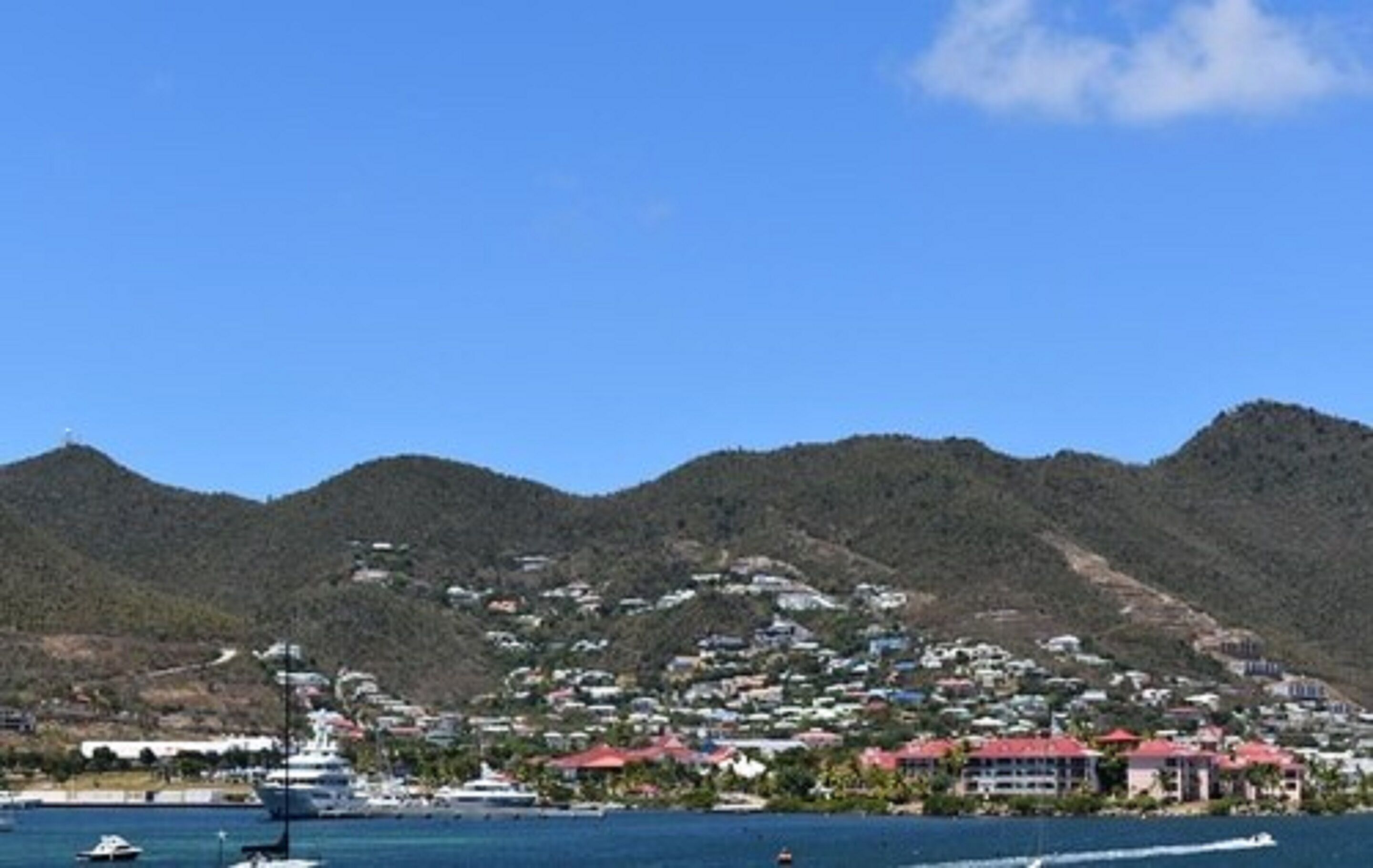 A coastal town with a marina, houses with red roofs, and hills in the background.